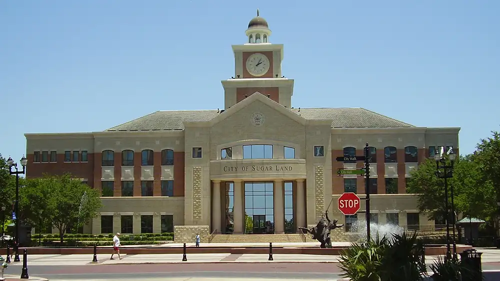 Exterior of Sugar Land City Hall with a central clock tower, arched windows and a fountain and stop sign in the foreground. Photo: WhisperToMe (public domain).
