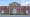 Front view of a two-story red brick courthouse with white trim, a central peaked roof, three white entrances and access ramps across the front.